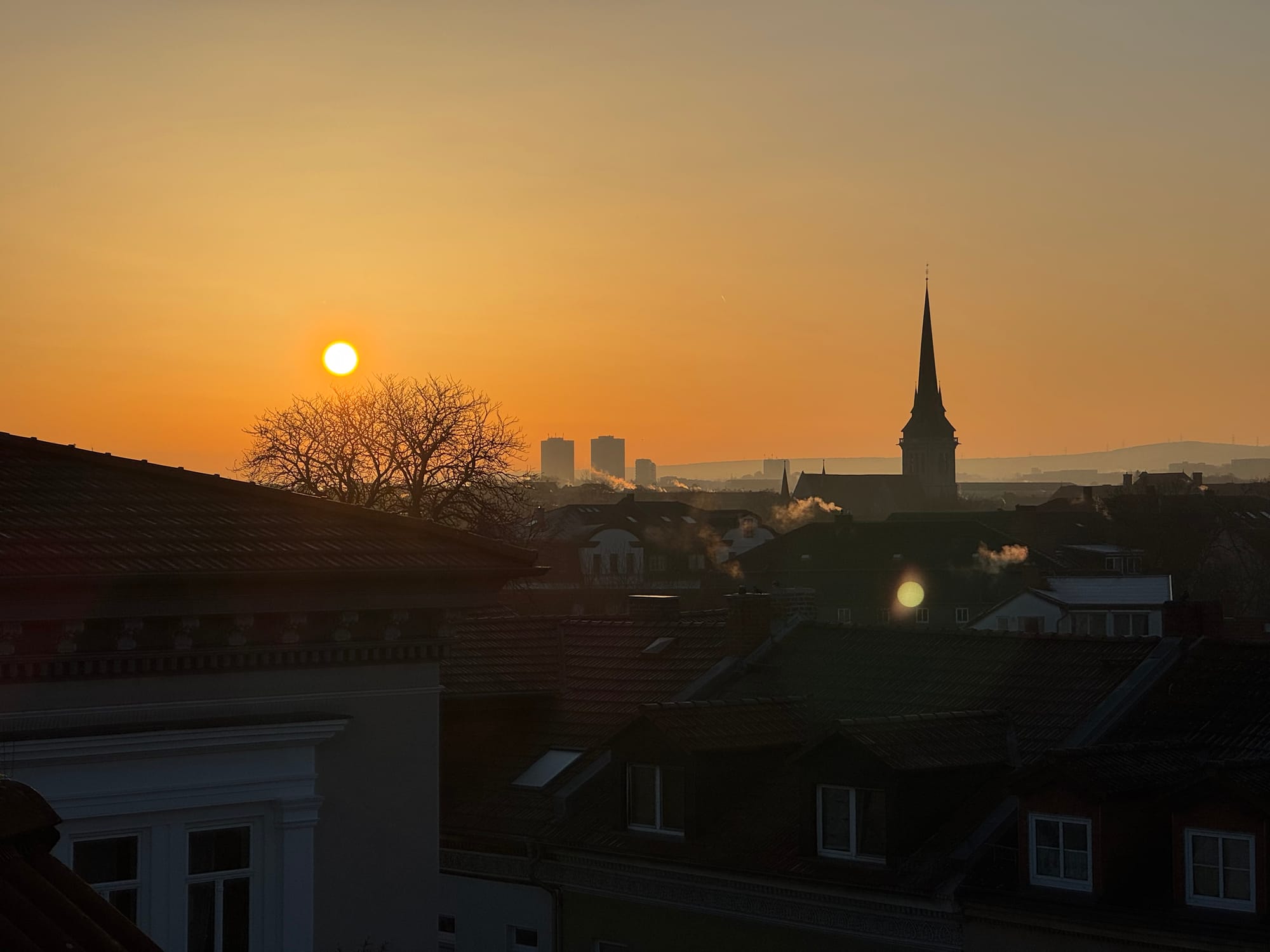 Die Sonne geht über  Stadt auf, der Himmel ist rot und eine kirchturmspitze ragt in den wolkenlosen Himmel.